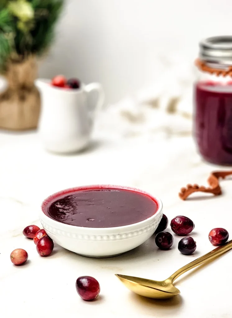 Smooth cranberry sauce in a white bowl with fresh cranberries and a gold spoon beside it.