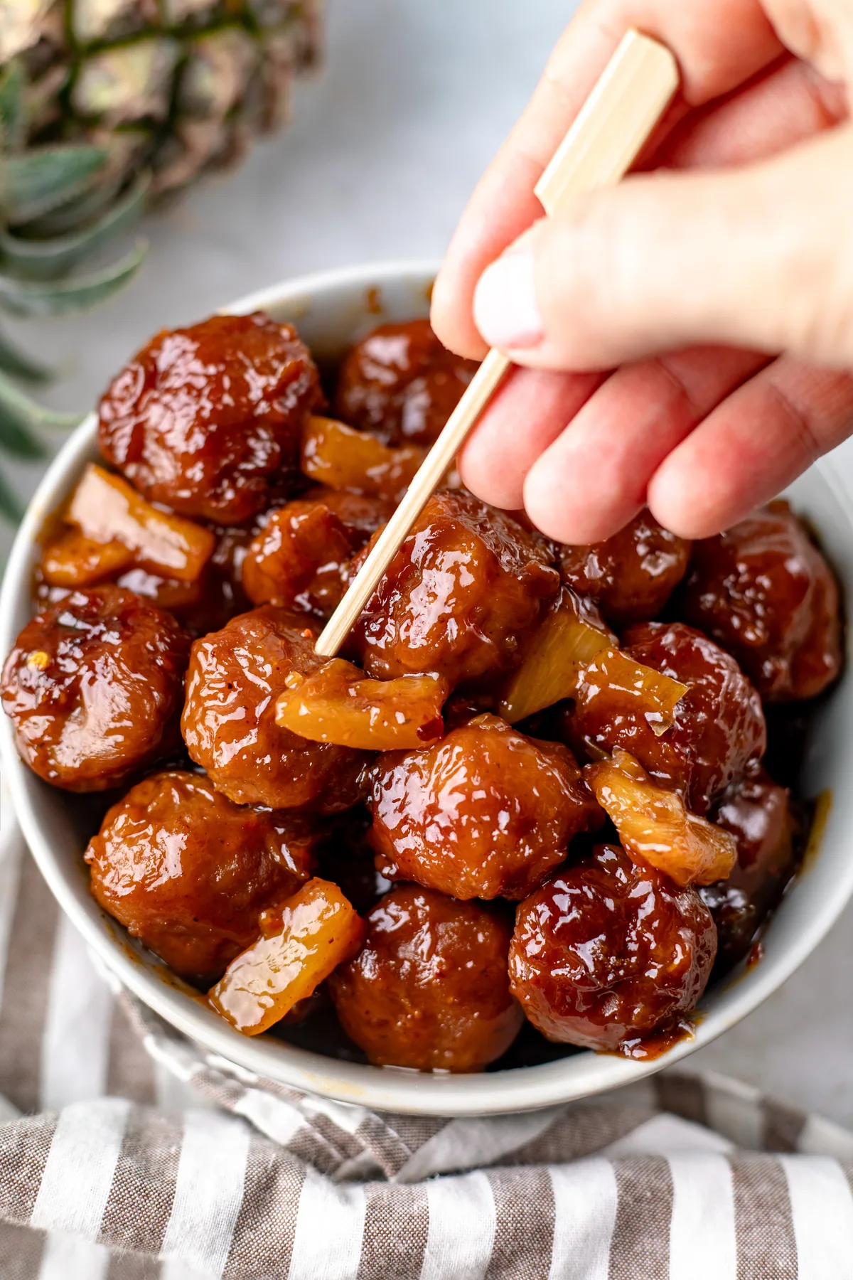 A hand sliding a Hawaiian meatball and a pineapple chunk onto a bamboo pick from a white bowl