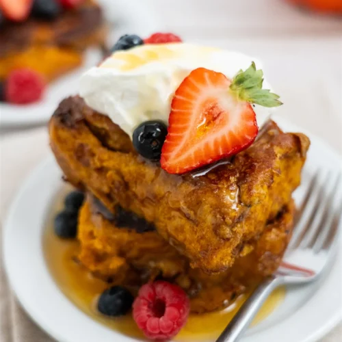 Two pieces of pumpkin cinnamon roll French toast stacked on a plate with whipped cream, a strawberry, berries and maple syrup