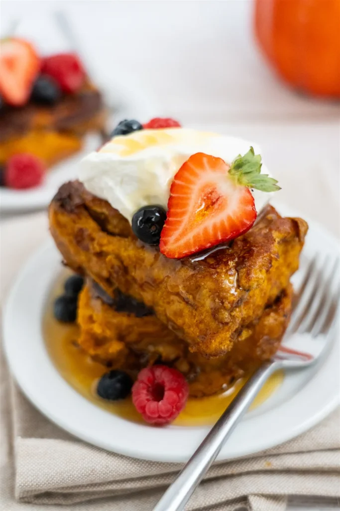 Two pieces of pumpkin cinnamon roll French toast stacked on a plate with whipped cream, a strawberry, berries and maple syrup