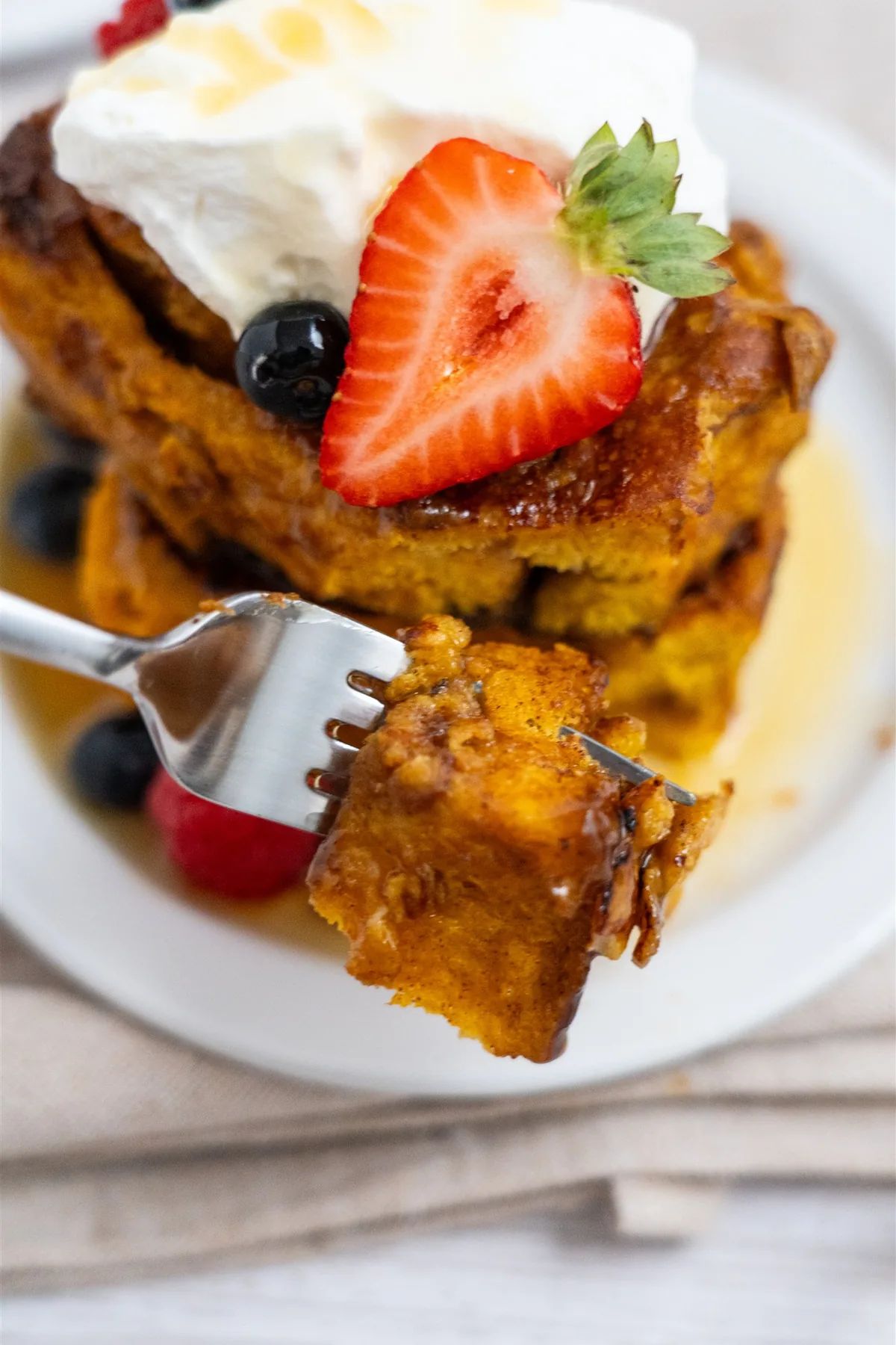 Fork holding a bite of pumpkin cinnamon roll French toast in front of a stack topped with whipped cream and a strawberry