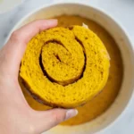 Hand holding a pumpkin cinnamon roll half with the swirl showing, over a bowl of pumpkin custard