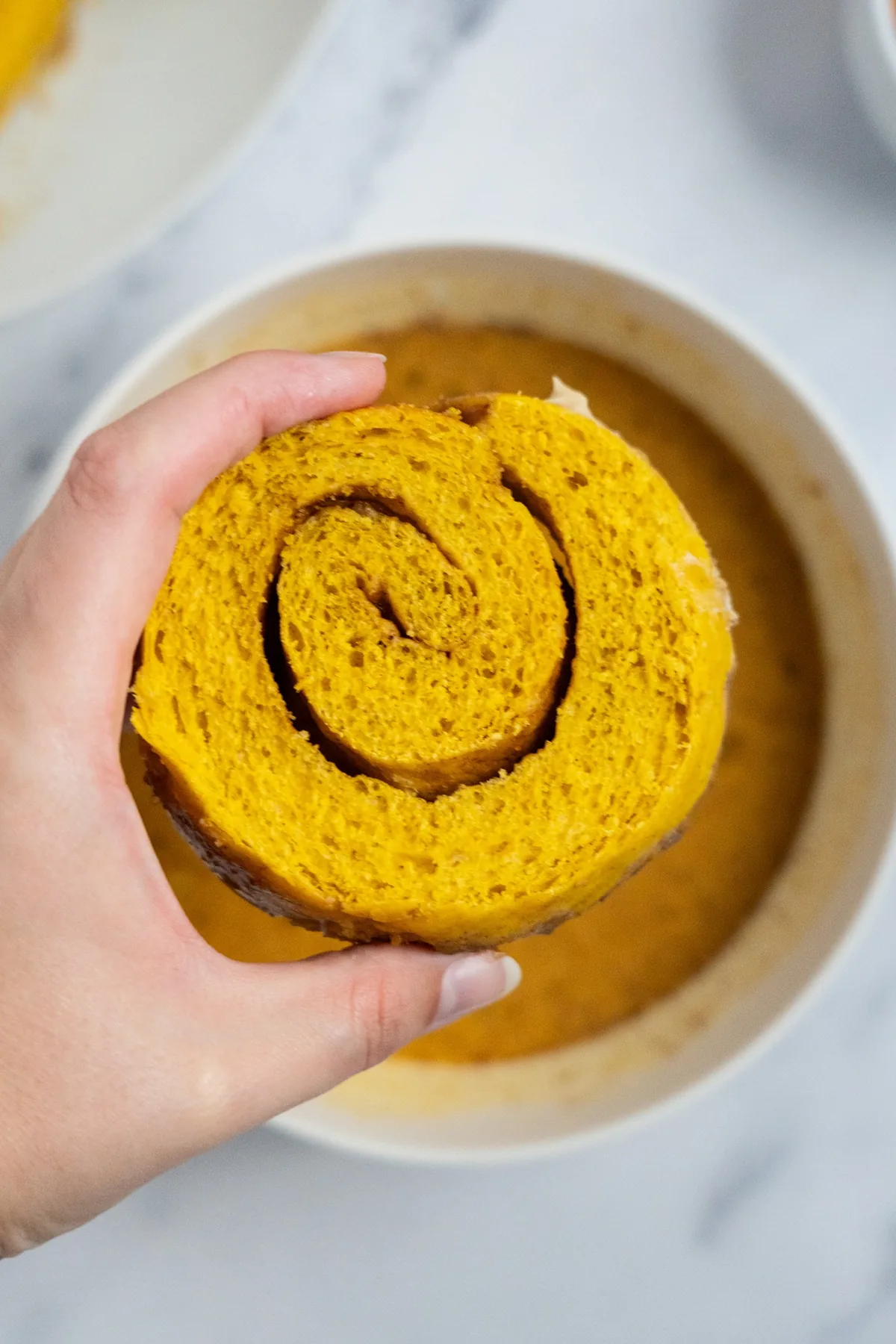Hand holding a pumpkin cinnamon roll half with the swirl showing, over a bowl of pumpkin custard