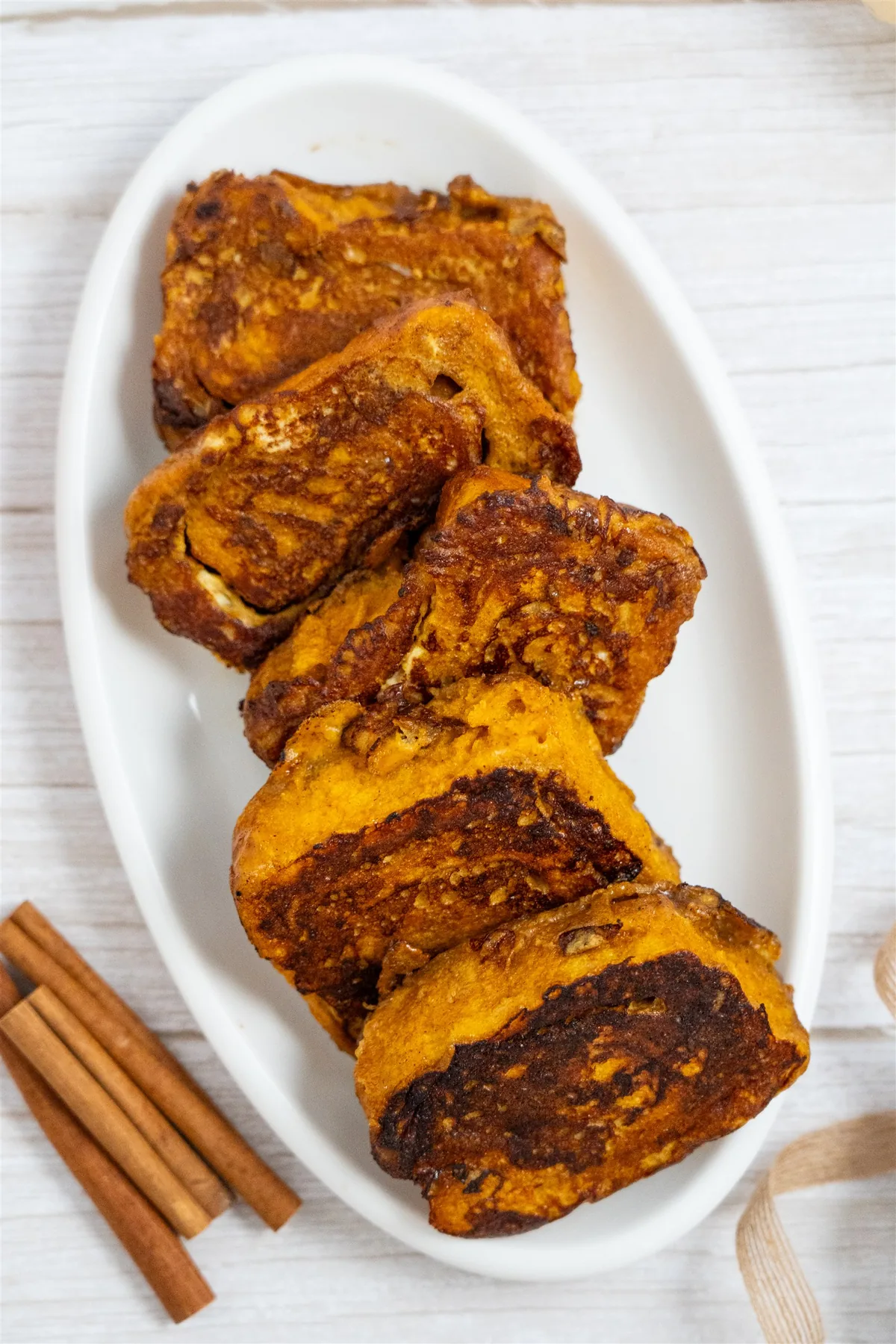 Pumpkin cinnamon roll French toast pieces with dark caramelized edges on a white oval platter with cinnamon sticks
