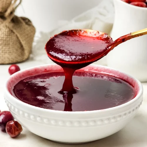 Smooth cranberry sauce flowing from a gold spoon into a white bowl.