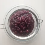 Warm cooked cranberry sauce being pressed through a fine-mesh sieve over a glass bowl.