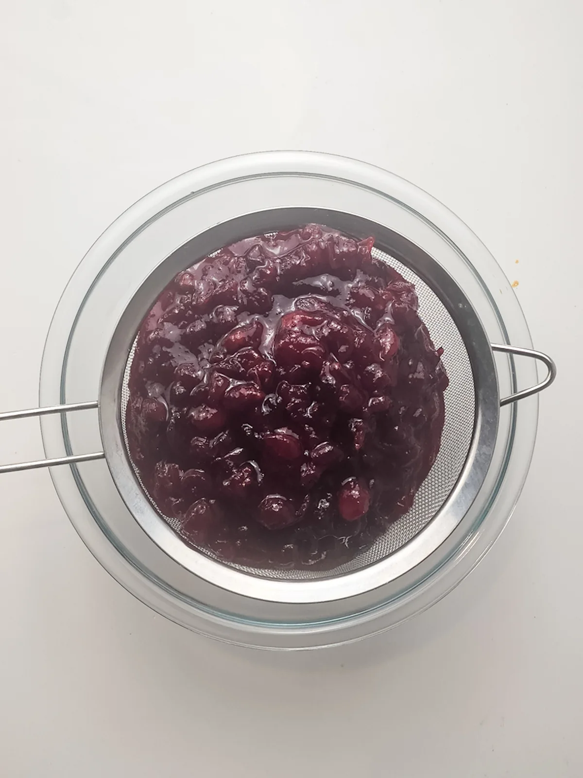 Warm cooked cranberry sauce being pressed through a fine-mesh sieve over a glass bowl.